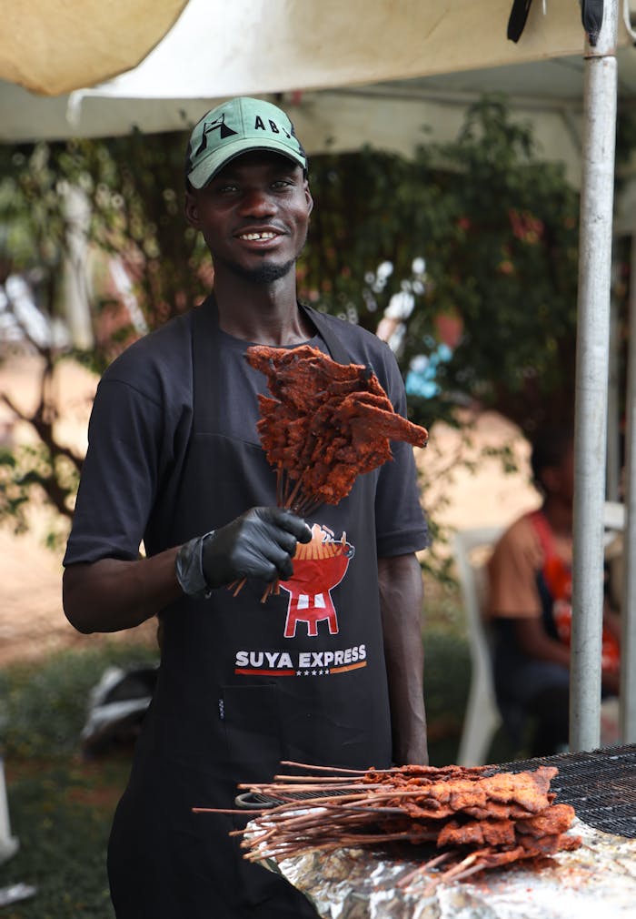 Man grilling traditional suya skewers outdoors at a food stall, smiling and serving customers.
